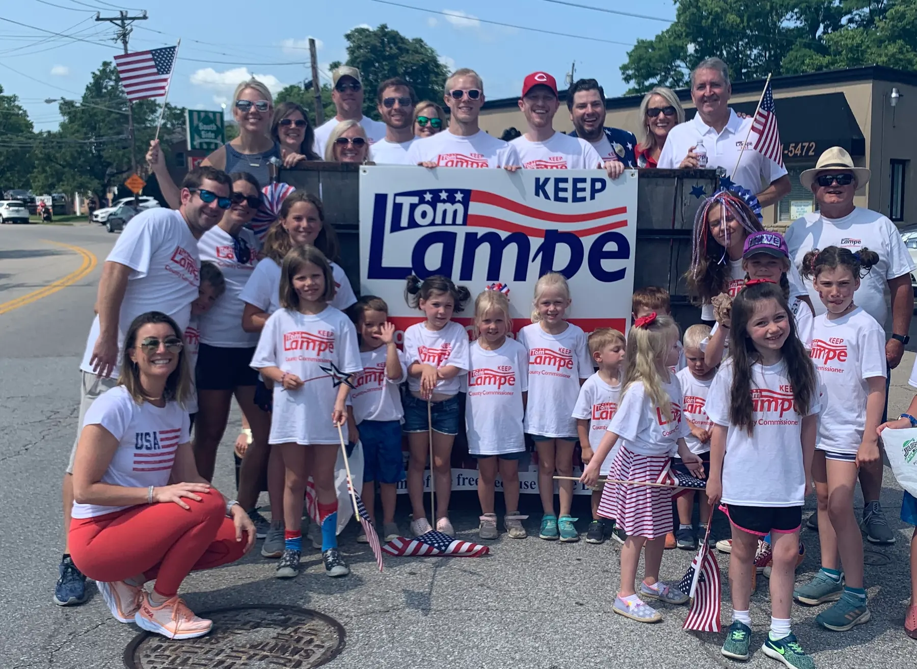 Tom Lampe stands with neighbors, parents and children in Tom Lampe T-shirts. They are next to a Keep Tom Lampe sign at a Campbell County parade.