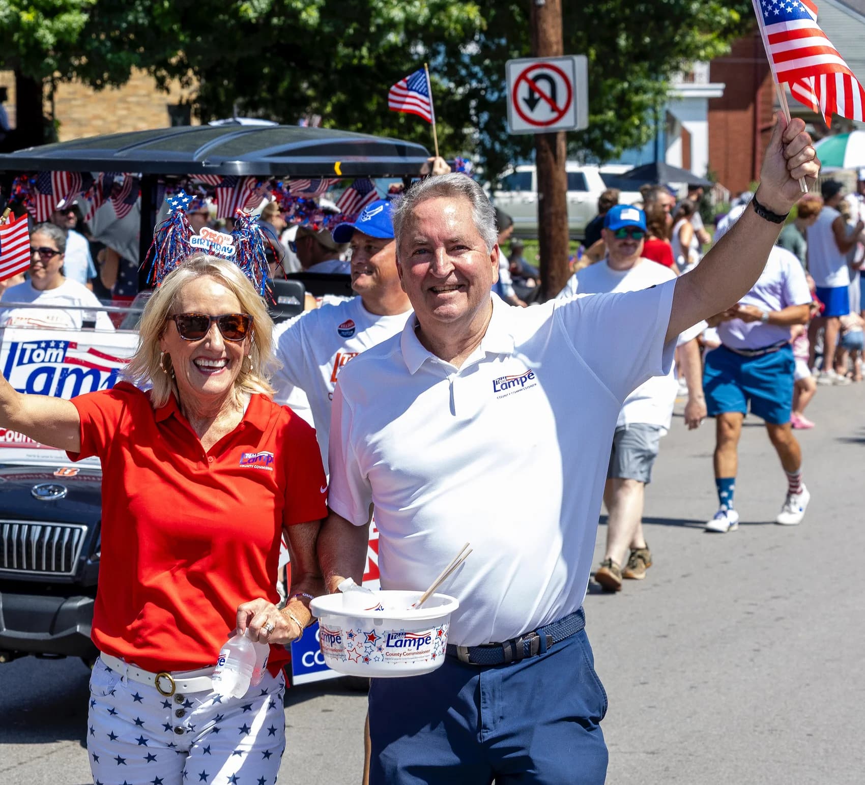 Tom Lampe marches in a Campbell County parade. He waves an American flag next to a woman in a red campaign shirt. Tom Lampe yard signs and neighbors line the street.